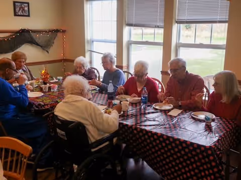 A group of elderly people sitting around a table covered with a black and red polka dot tablecloth, eating and drinking in a well-lit room with large windows. The room has light-colored walls and some decorations, including a small string of lights and a framed picture on the wall.