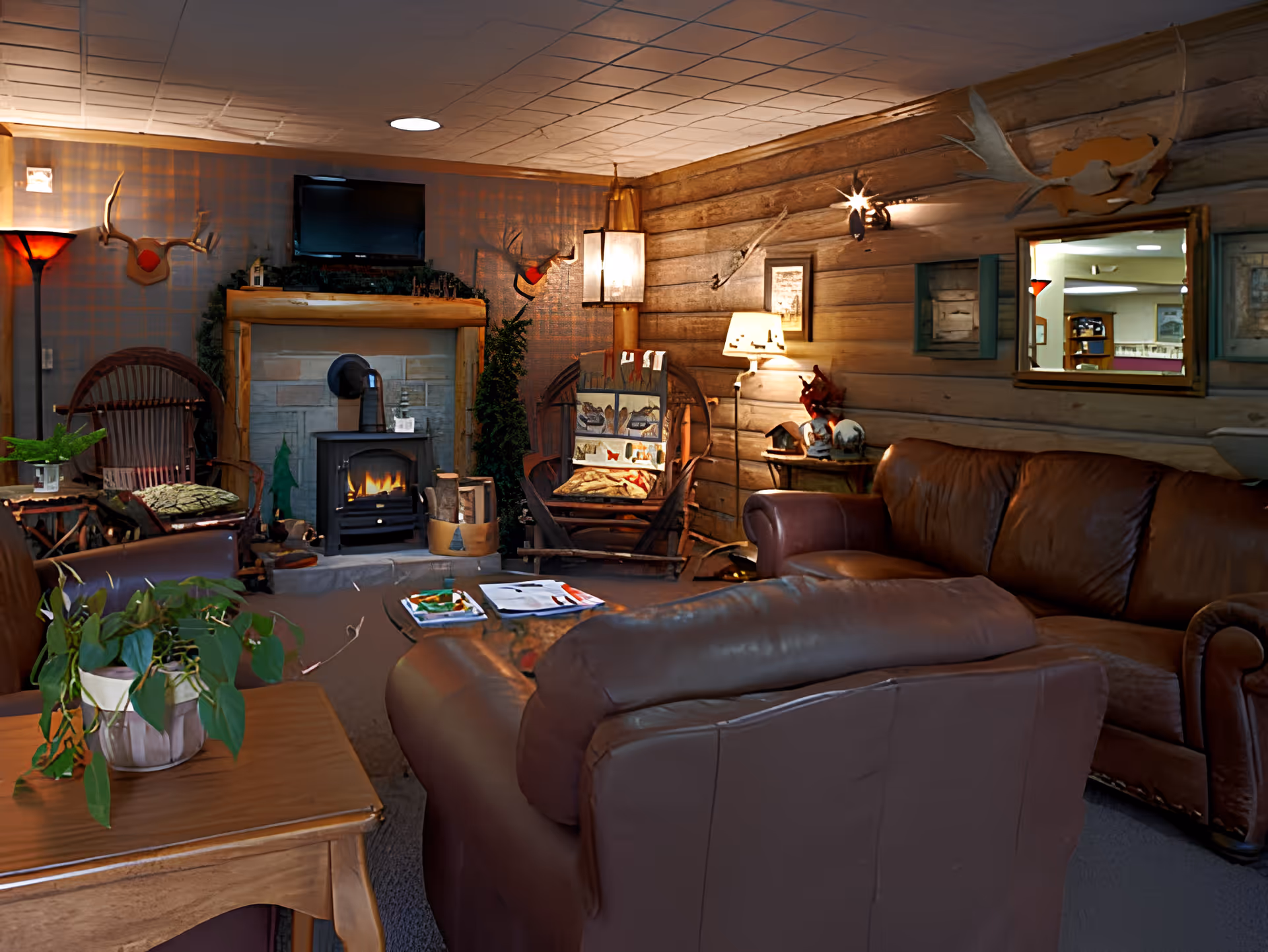 Cozy living room with brown leather sofas and wooden chairs arranged around a small fireplace with a TV mounted above it. The walls are wooden logs decorated with mounted antlers and a mirror. A wooden table with a potted plant and magazines is in the foreground, and warm lighting creates a comfortable atmosphere.