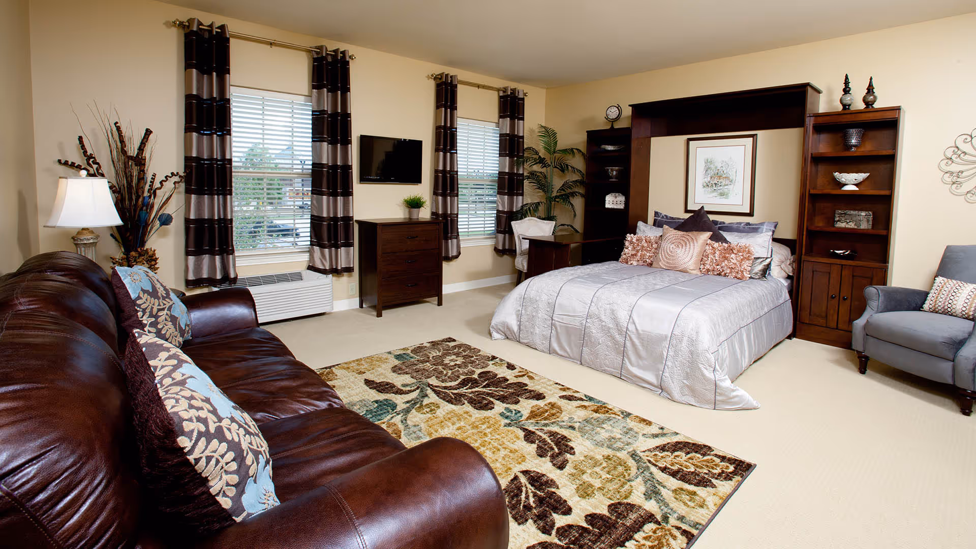 A cozy bedroom featuring a large bed with multiple decorative pillows, a dark wood headboard with built-in shelves, a patterned area rug, a brown leather sofa with floral pillows, two windows with striped curtains, a wall-mounted TV, a small dresser, a gray armchair, and decorative plants and artwork on the walls.