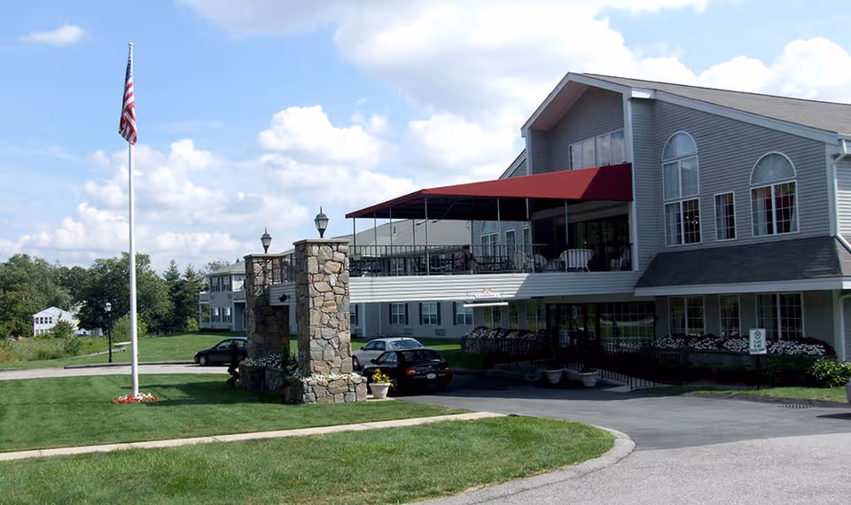 Exterior view of a senior living facility building with a stone entrance structure, an American flag on a flagpole, a covered balcony with a red awning, parked cars, and well-maintained green lawns under a partly cloudy sky.