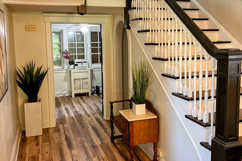 Interior hallway of a senior living facility with wooden flooring, a staircase with white spindles and dark handrail, a small wooden table with a potted plant, and another potted plant in a tall white planter. In the background, a room with windows, a white cabinet, and a purple orchid is visible.