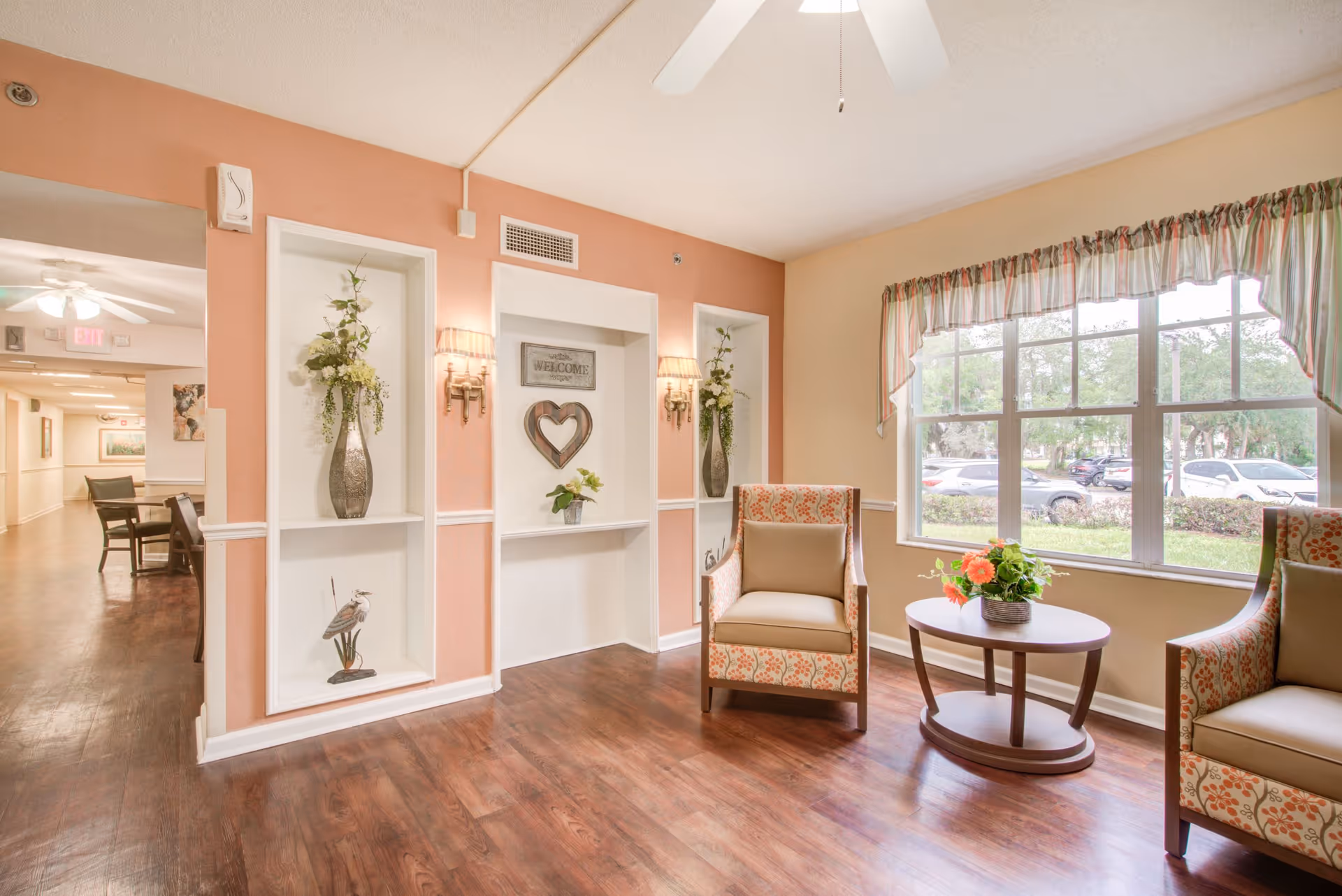 A bright and welcoming sitting area in a senior living facility with two patterned armchairs and a round wooden table with a flower arrangement. The room has large windows with striped valances letting in natural light, wooden flooring, and a peach-colored accent wall with built-in shelves decorated with vases, flowers, and a heart-shaped wall hanging. A hallway with more seating and artwork is visible in the background.