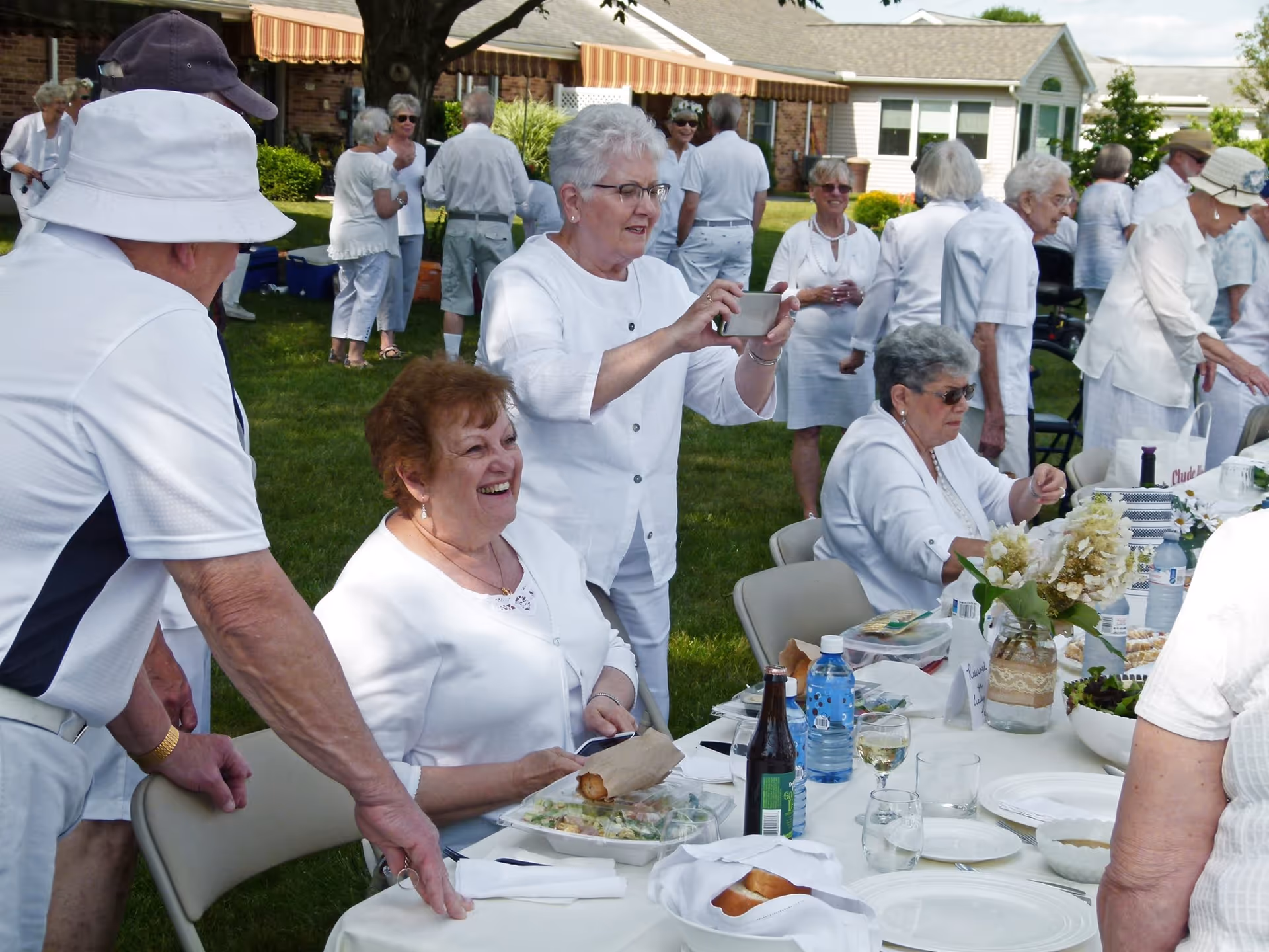A group of elderly people dressed mostly in white are gathered outdoors on a lawn for a social event. Some are seated at a long table with food, drinks, and floral centerpieces, while others stand and mingle in the background. One woman is taking a photo with a smartphone. The setting appears to be a garden or patio area of a residential facility.