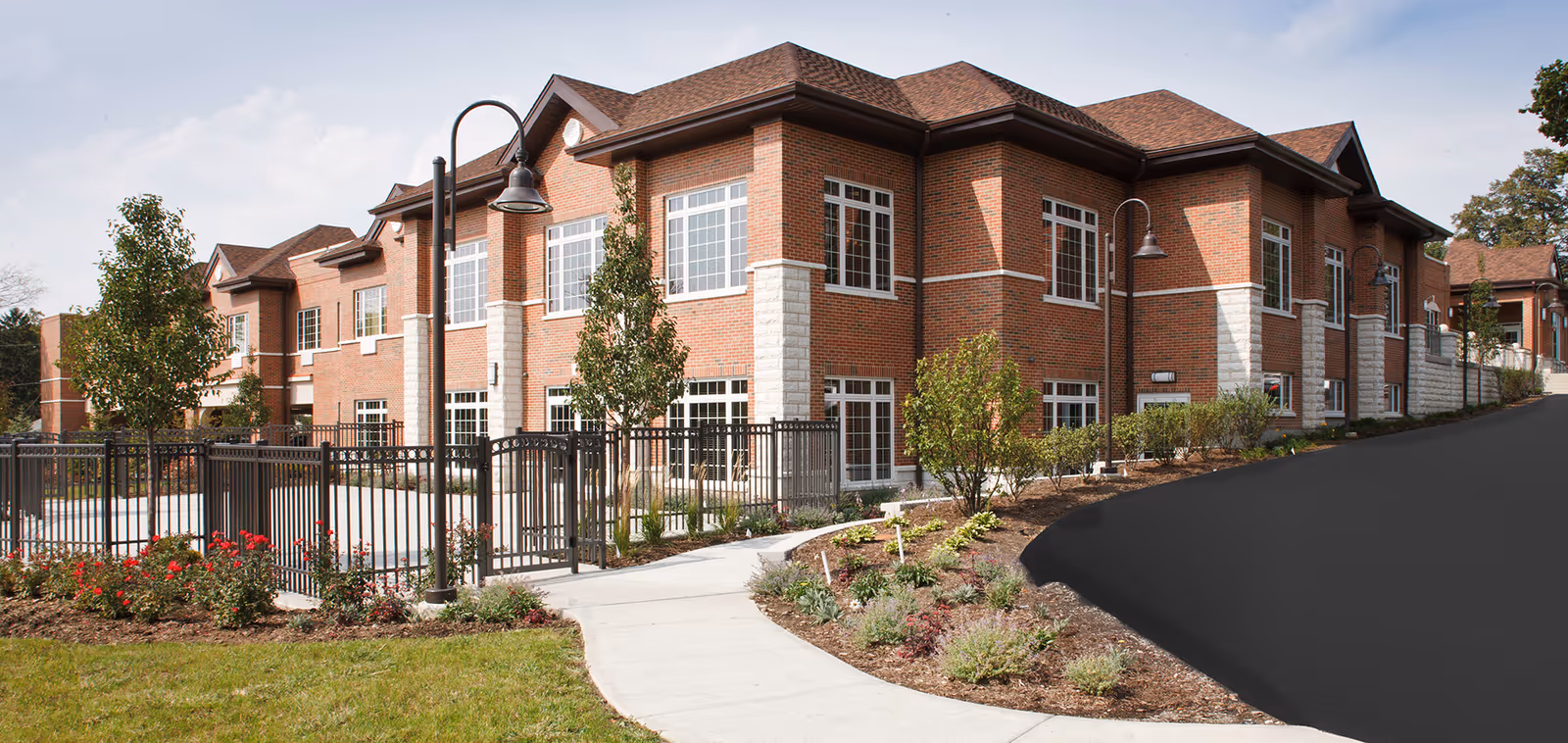 Exterior view of a two-story brick building with large windows, surrounded by a landscaped garden with bushes, flowers, and trees. A black metal fence and street lamps line the walkway leading to the entrance.