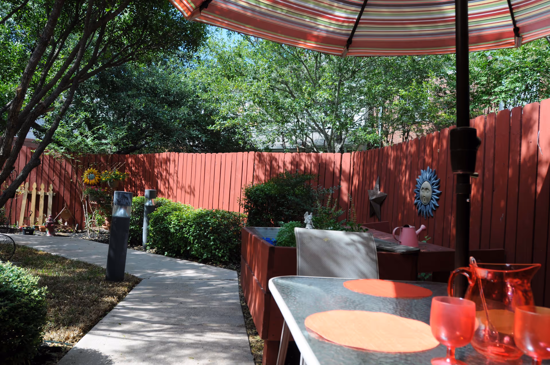 Outdoor patio area with a glass table set with orange placemats and red glasses under a striped umbrella. The patio is surrounded by a red wooden fence with decorative items like a sun face and star hanging on it. There are green bushes and trees providing shade along a concrete pathway.
