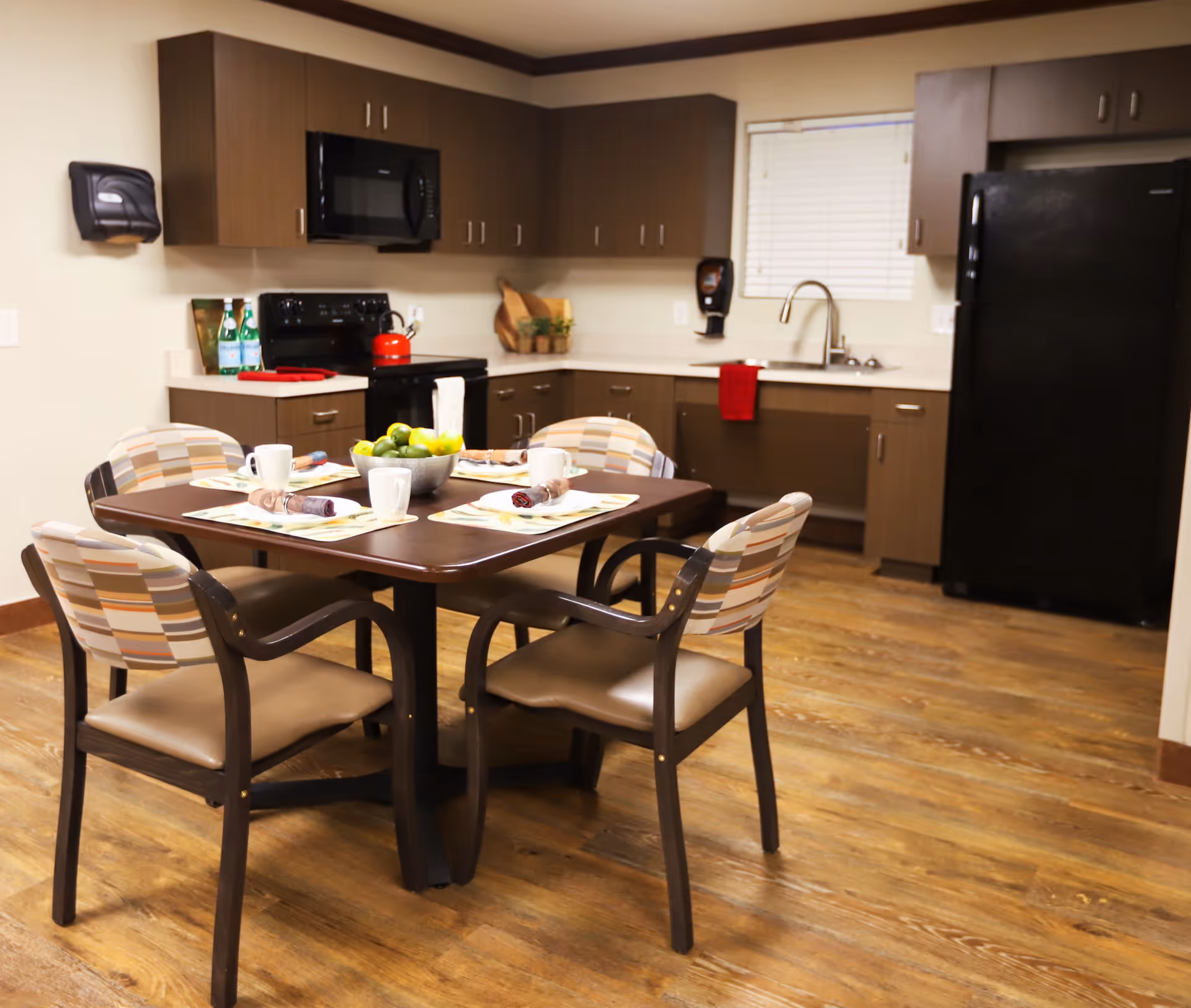 A modern kitchen and dining area with wooden cabinets, a black refrigerator, stove, and microwave. A dining table with four chairs is set with placemats, cups, and napkins. The floor has a wood finish, and there is a window with blinds above the sink.