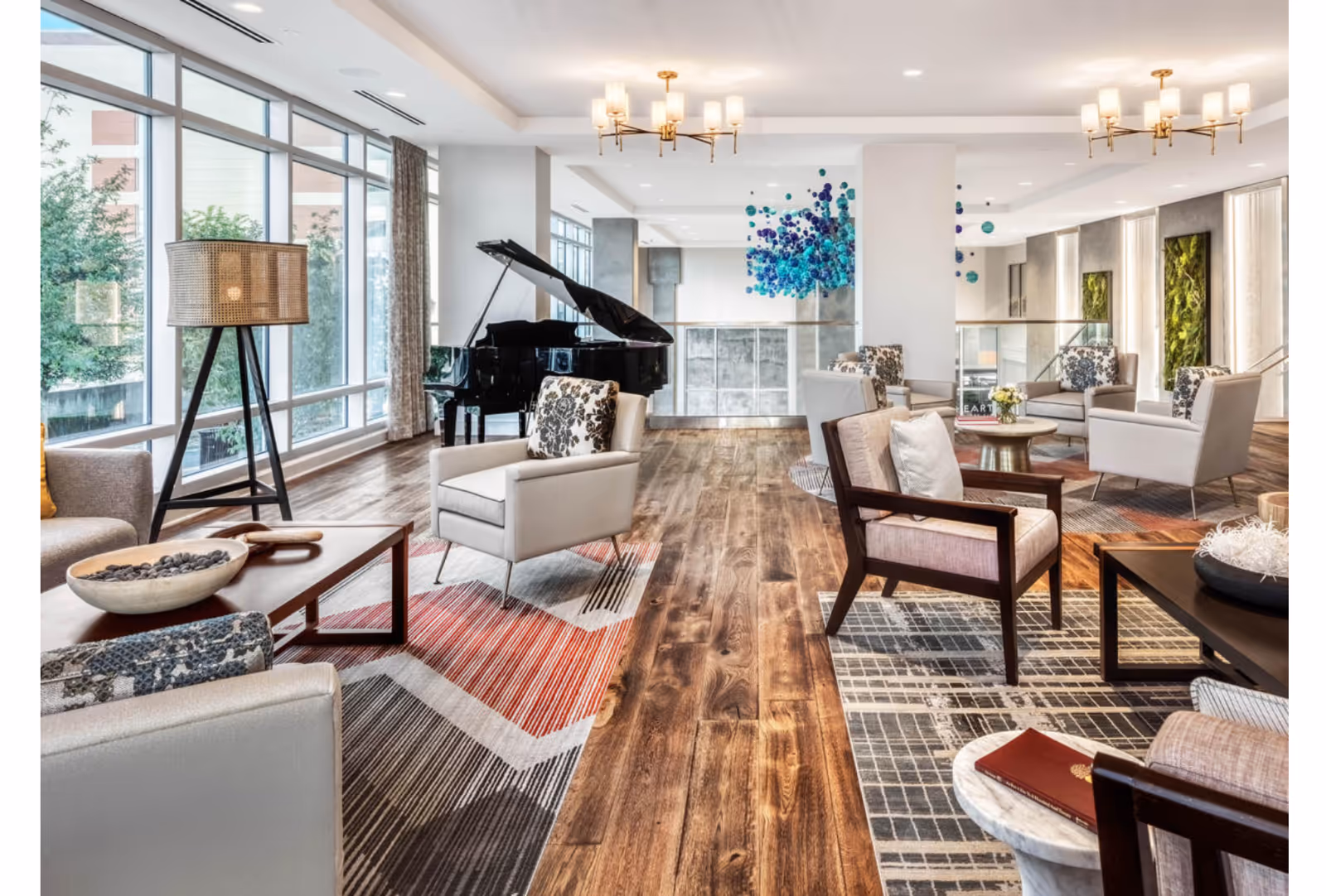 A spacious and well-lit senior living facility common area with large windows, wooden flooring, multiple armchairs with patterned cushions, coffee tables, a floor lamp, and a black grand piano. The room features modern chandeliers and decorative wall art with blue and green accents.