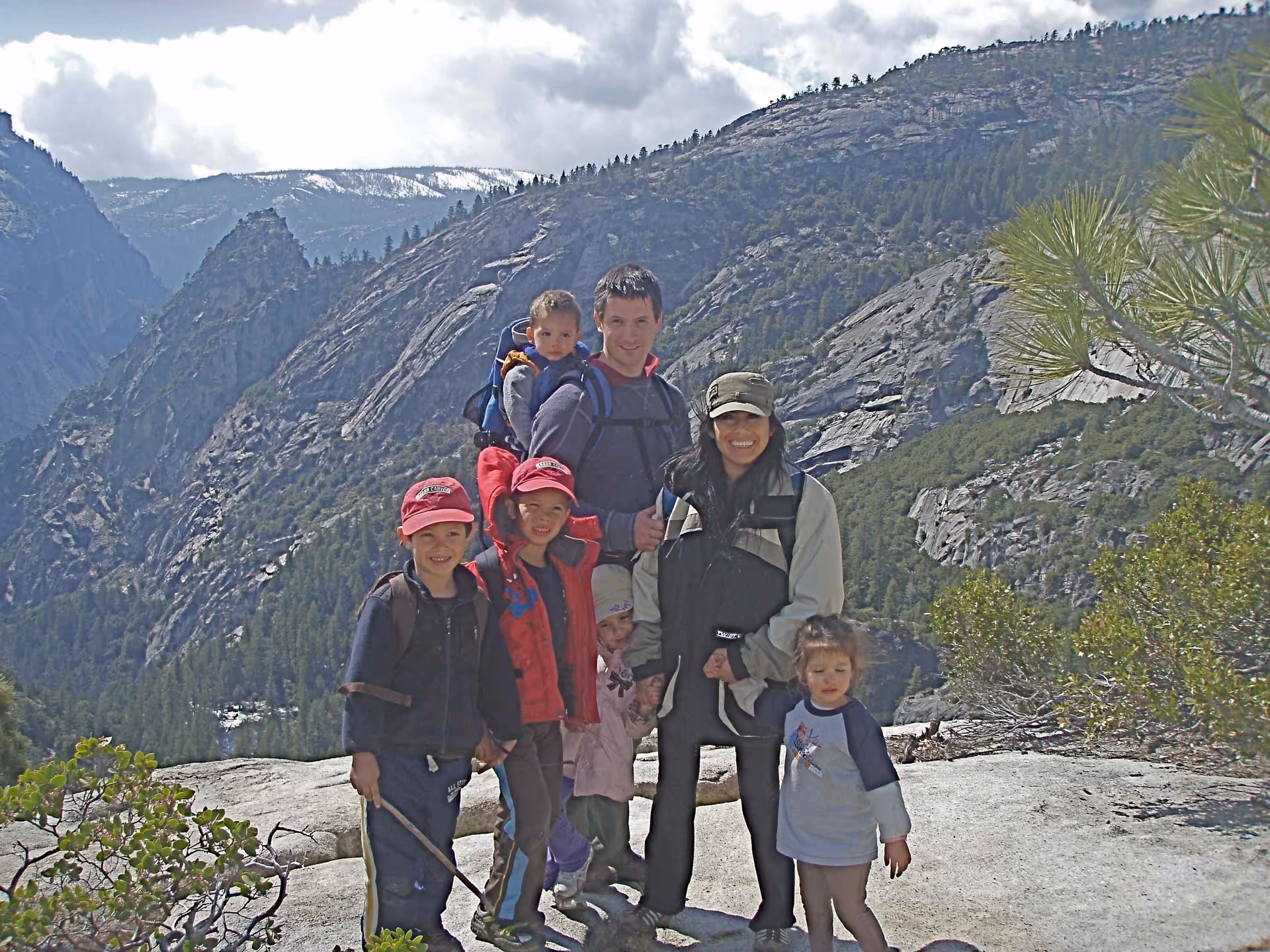 A group of six people, including two adults and four children, posing on a rocky mountain trail with forested hills and cloudy sky in the background.