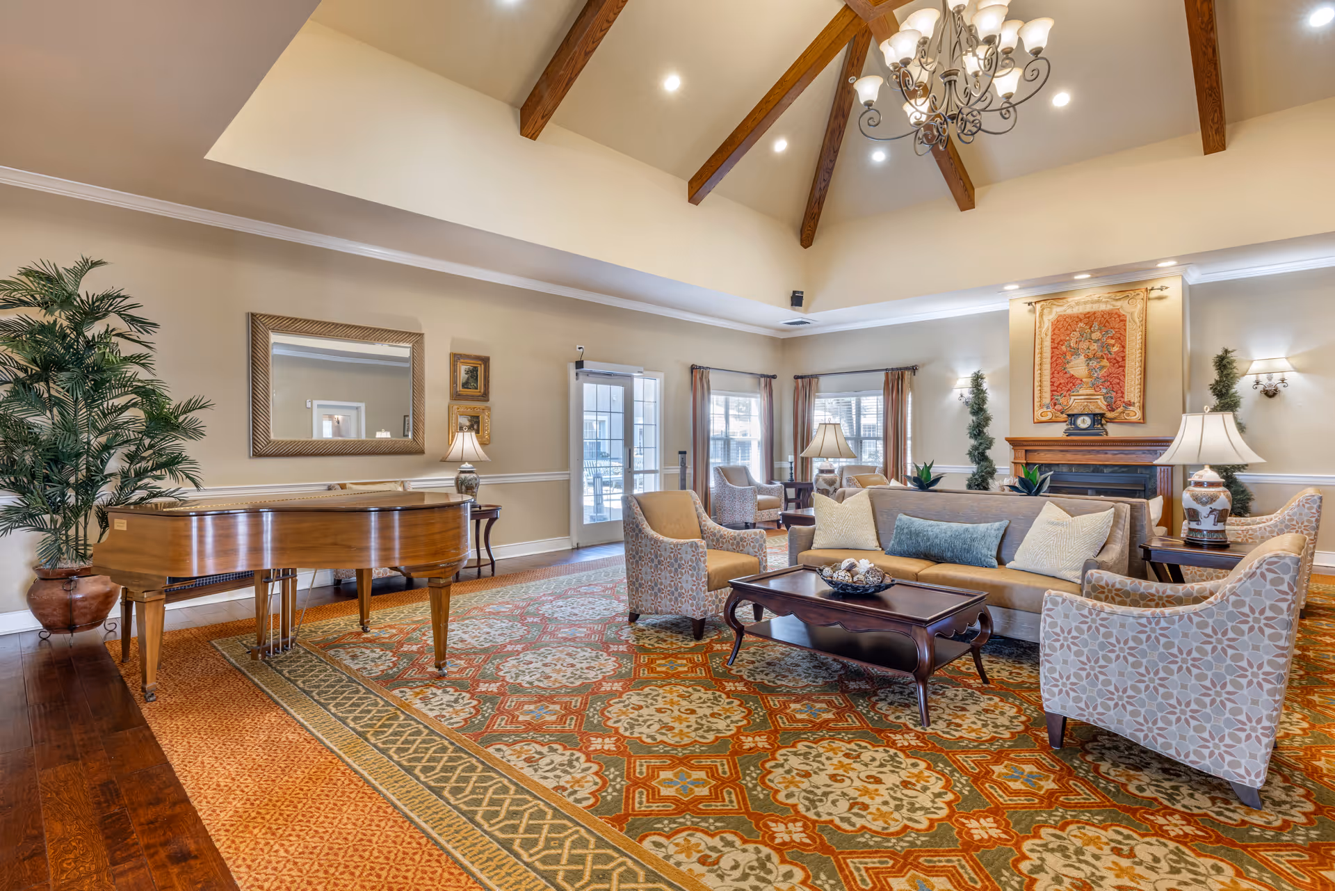 A spacious and elegant living room with a high vaulted ceiling featuring wooden beams and a chandelier. The room includes a grand piano, a large patterned area rug, a sofa with cushions, several armchairs, a coffee table, side tables with lamps, a fireplace with decorative artwork above it, and large windows with curtains allowing natural light to enter.