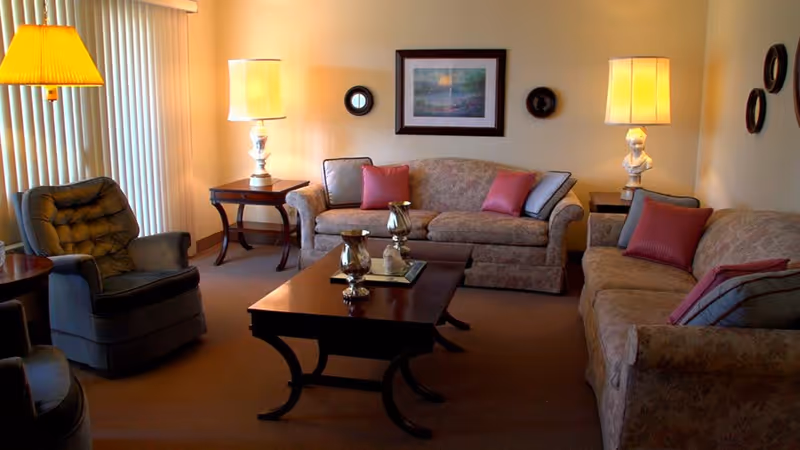A cozy living room with two floral-patterned sofas adorned with red and gray pillows, a dark wooden coffee table with decorative items, two side tables each with a lamp, a cushioned armchair, vertical blinds covering a window, and framed artwork on the wall.