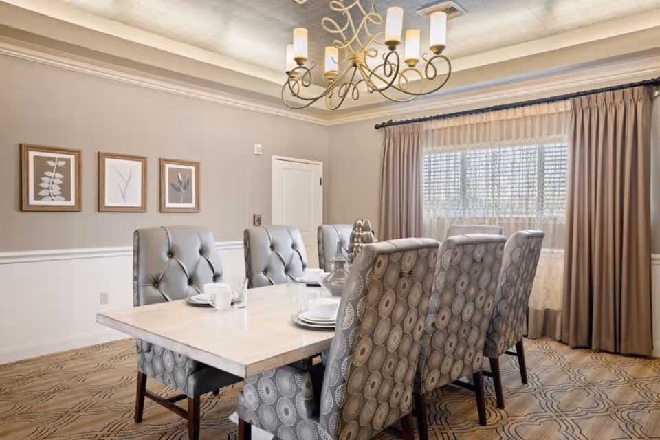 Dining room with a light wood table surrounded by tufted upholstered chairs, a chandelier overhead, and a window with curtains.