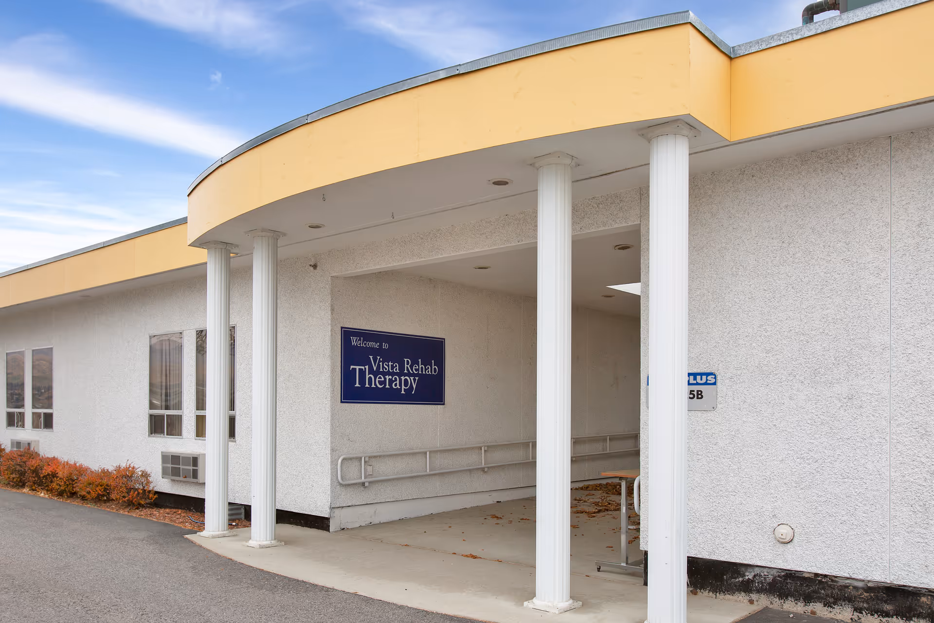 Exterior view of a building entrance with white columns and a yellow overhang. A blue sign on the wall reads 'Welcome to Vista Rehab Therapy'. There are windows and some bushes along the side of the building under a partly cloudy sky.