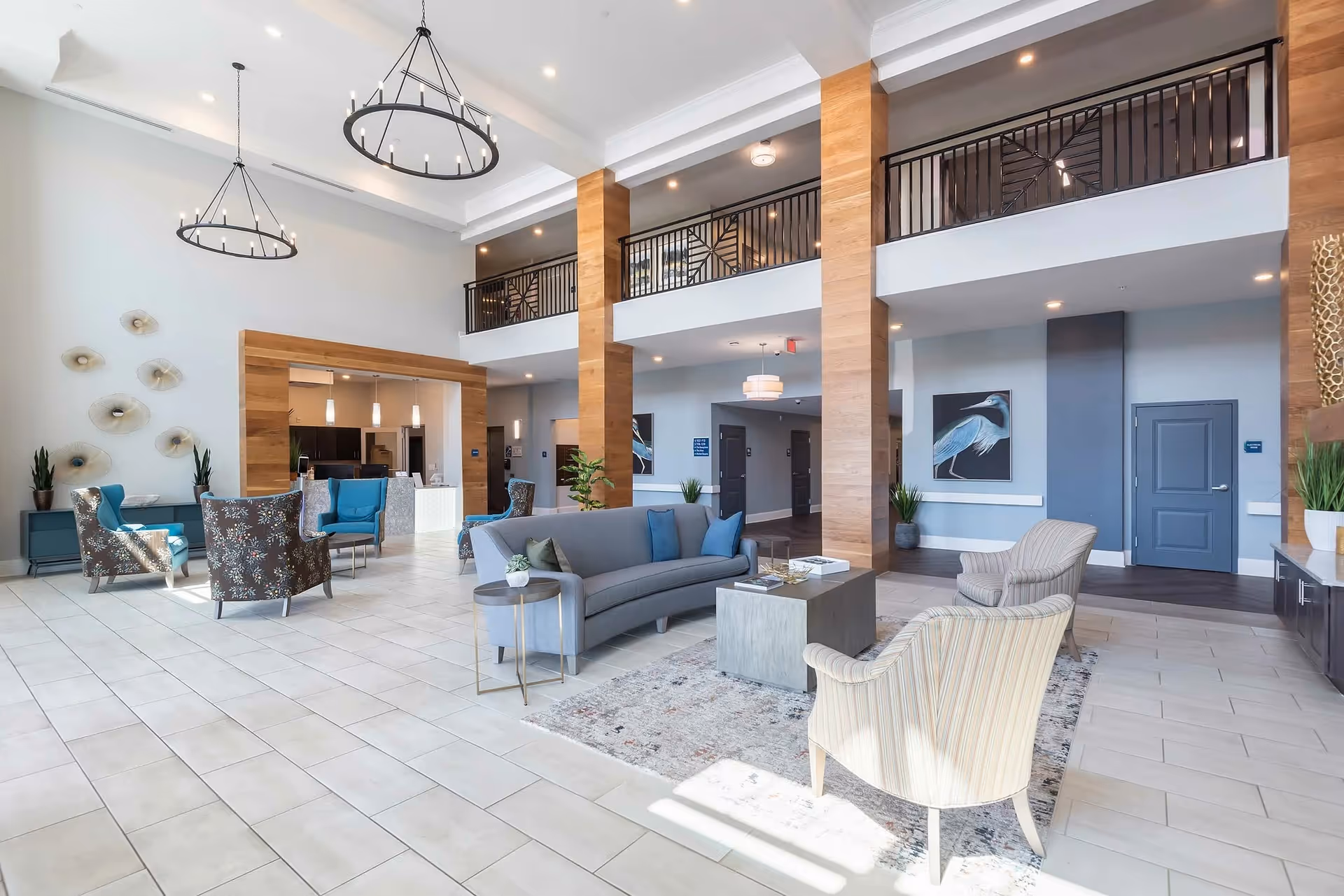 A spacious and modern senior living facility common area with high ceilings, large windows, and contemporary furniture including a curved gray sofa, patterned armchairs, and small tables. The room features wooden pillars, decorative wall art, and two large circular chandeliers hanging from the ceiling. There is a balcony railing on the upper level and a reception or kitchen area in the background.