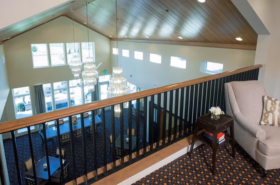 View from an upper level overlooking a dining area with multiple tables and chairs. The space features large windows letting in natural light, three hanging chandeliers, and a high wooden ceiling. In the foreground, there is a cozy armchair with a patterned pillow and a small side table holding a small flower arrangement and some books.