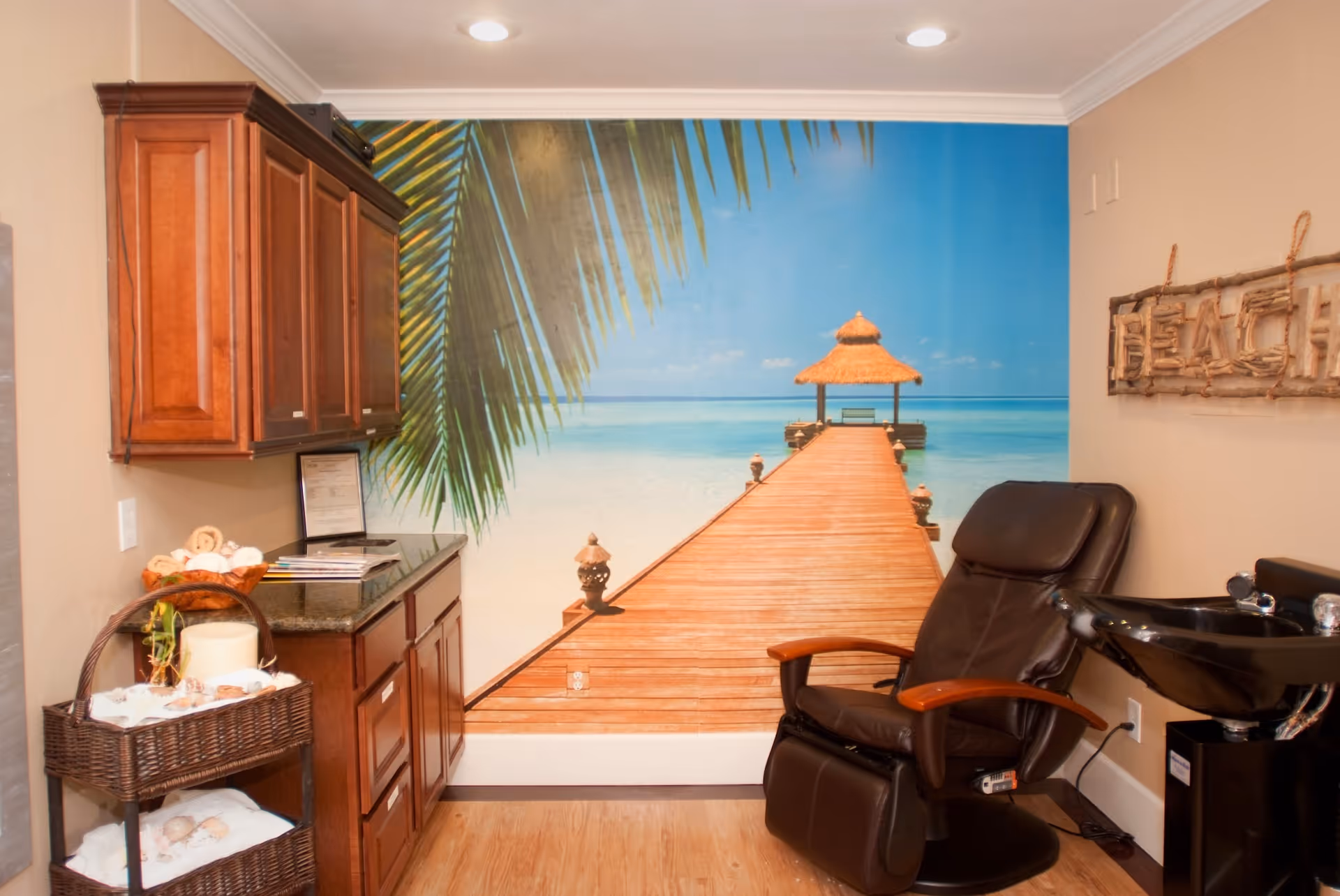 A cozy salon room with a black reclining chair next to a black hair washing sink. The room features wooden cabinets and a wicker basket with towels and spa items. The back wall has a large mural of a wooden pier extending into a tropical beach with clear blue water and a thatched gazebo. A decorative sign that reads 'BEACH' is mounted on the adjacent wall.