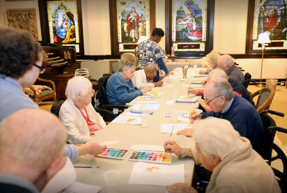 A group of elderly individuals seated around a long table engaged in a painting activity with watercolor palettes and brushes. A caregiver stands nearby assisting. The room features stained glass windows and warm lighting.