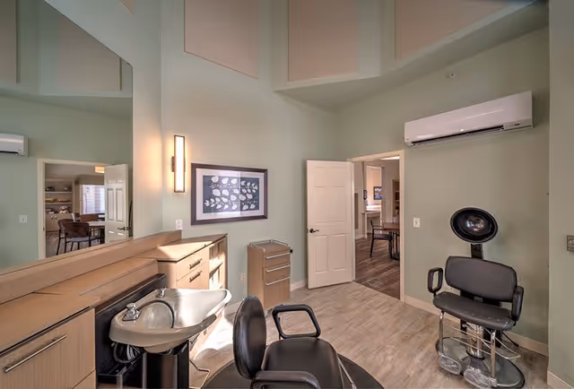 Interior view of a senior living facility room with a hair washing station, including a sink with a faucet, a black salon chair, and a black hair dryer chair. The room has light green walls, a large mirror, a framed picture on the wall, and an open door leading to another room with wooden flooring and furniture. An air conditioning unit is mounted on the wall above the door.