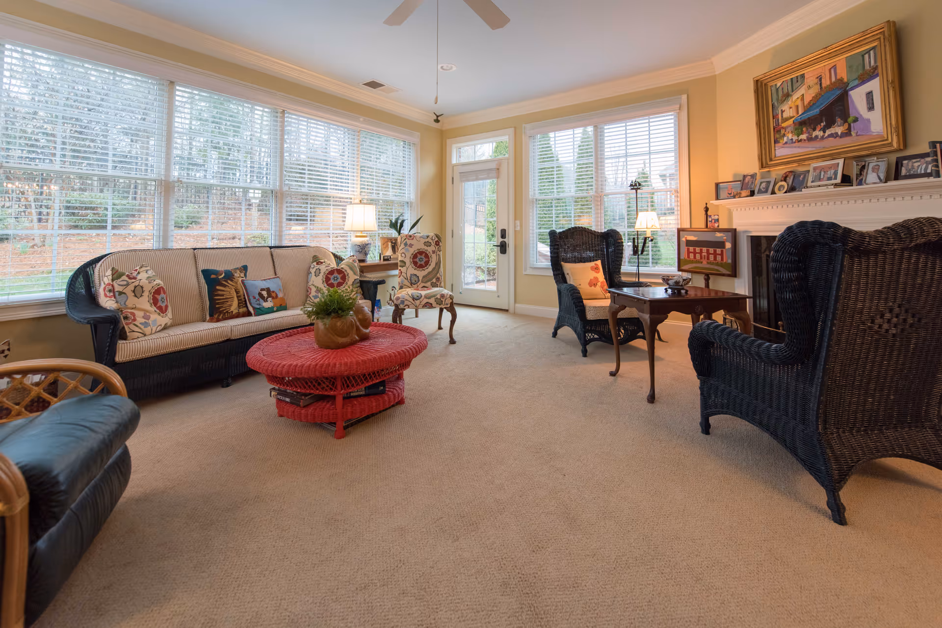 A cozy living room with large windows letting in natural light. The room features a beige carpet, a black wicker sofa with patterned cushions, two black wicker armchairs, a floral upholstered chair, a red round wicker coffee table with a plant on top, and a wooden side table with a lamp. There is a fireplace with framed photos and a colorful painting above it. A door leads outside to a garden area.