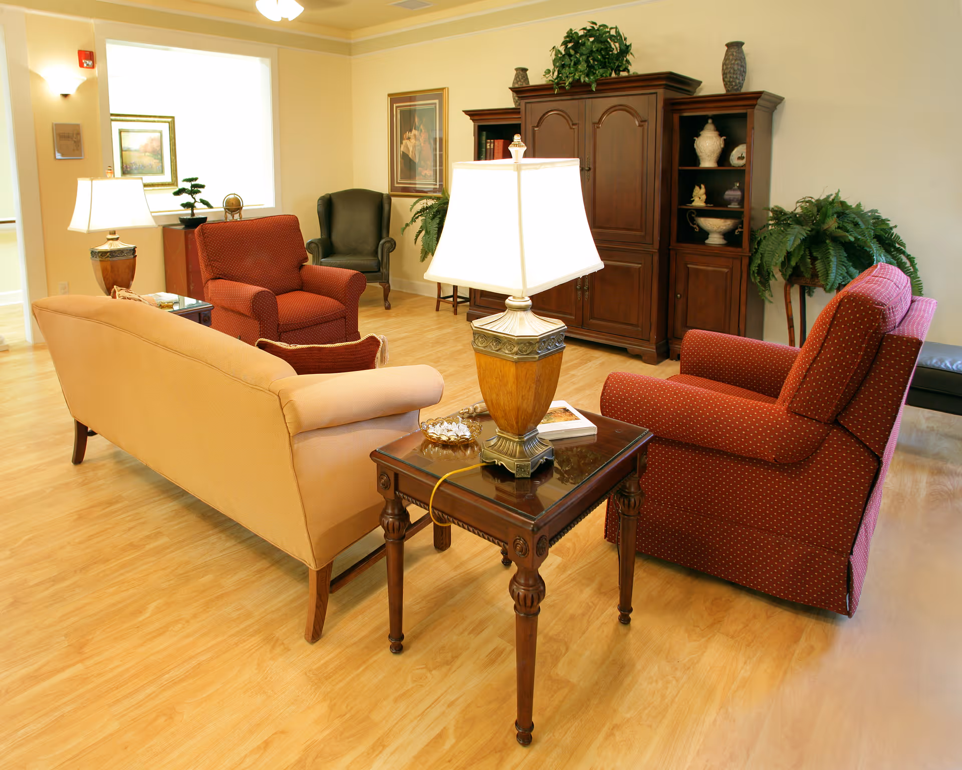 A cozy senior living facility common area with a beige sofa, two red armchairs, a green armchair, a wooden side table with a lamp, a large wooden cabinet, framed artwork on the walls, and potted plants.