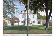 Front exterior view of a large white multi-story house with a manicured lawn, trees, and a black metal fence in front. The house has multiple windows and a small porch area.