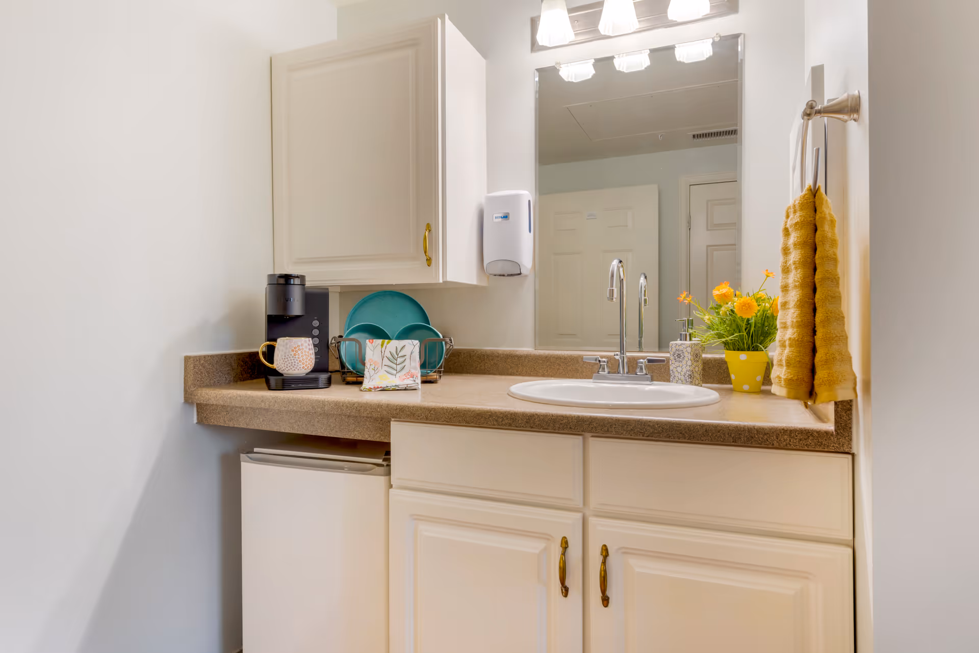 Vanity-style sink and mirror with a countertop coffee maker, mini refrigerator, yellow towels, and a small vase of flowers.