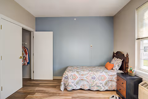 A bedroom with a single bed against a blue accent wall, featuring a patterned bedspread and an orange heart-shaped pillow. There is a wooden nightstand with drawers next to the bed, holding a few items. A window with blinds is on the right side, and an open closet with clothes hanging is on the left. The floor is wood laminate.