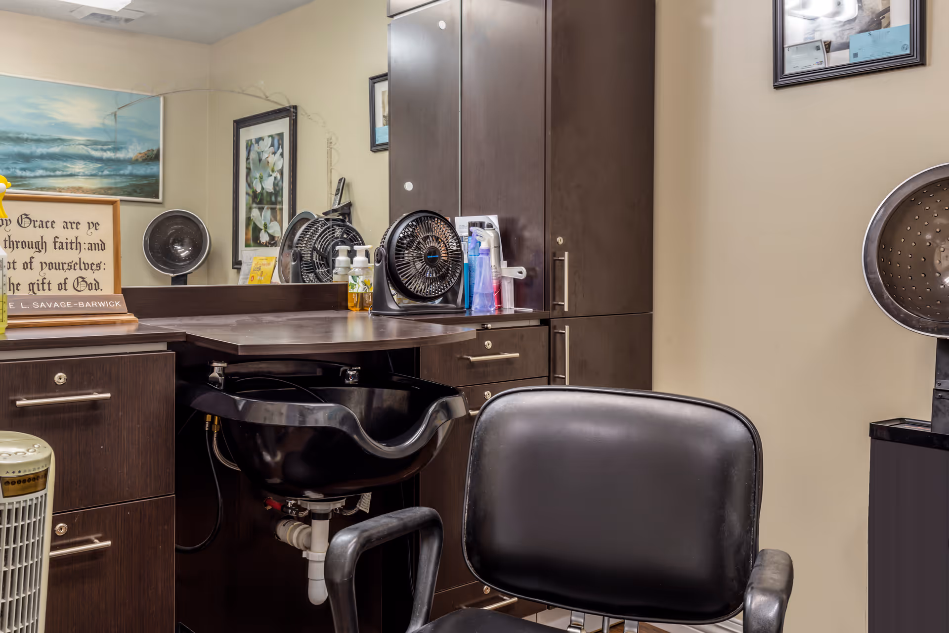 Interior hair salon station with a black styling chair, shampoo basin, cabinets, and hair-drying equipment.