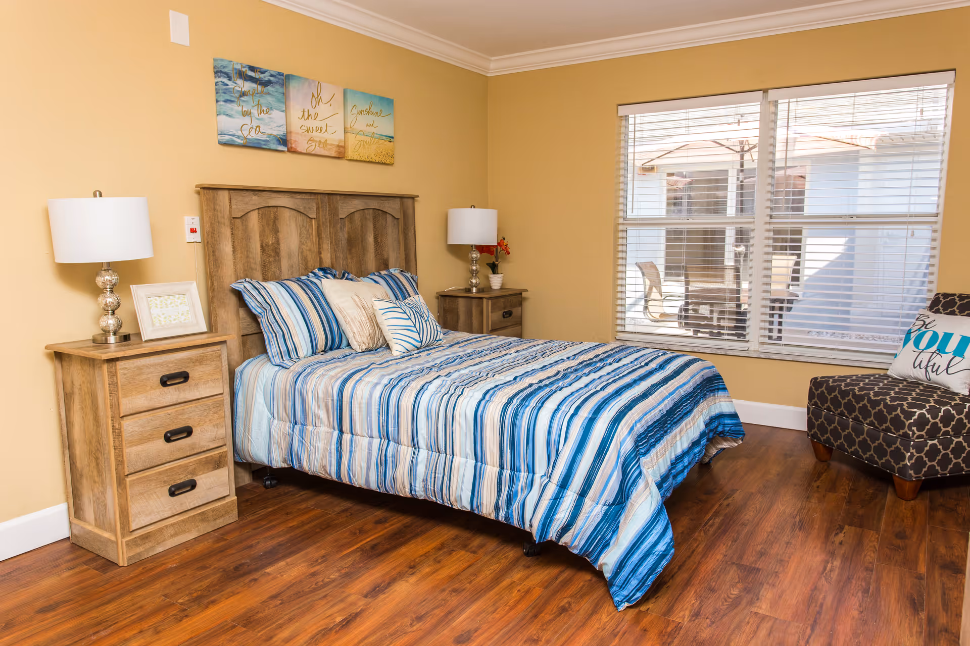 A cozy bedroom with a wooden bed frame and matching nightstands on either side. The bed is made with a blue and white striped comforter and several pillows. Two lamps with white shades sit on the nightstands. Above the bed are three decorative wall art pieces with beach themes and inspirational text. A window with blinds lets in natural light, and a patterned armchair with a decorative pillow is placed near the window. The room has wooden flooring and beige walls.