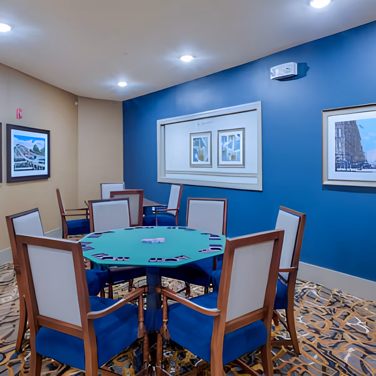 A small room with a round table covered with a green felt surface, surrounded by eight wooden chairs with blue cushions and white backs. The walls are painted blue and beige, decorated with framed artwork. The floor has a patterned carpet, and the ceiling has recessed lighting.