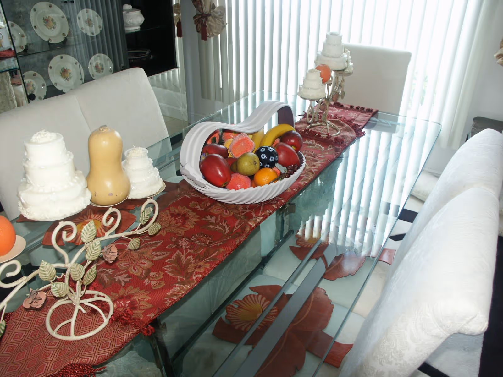 Glass dining table with a red table runner, decorative fruit bowl and faux cakes, surrounded by upholstered chairs in a bright room with vertical blinds.