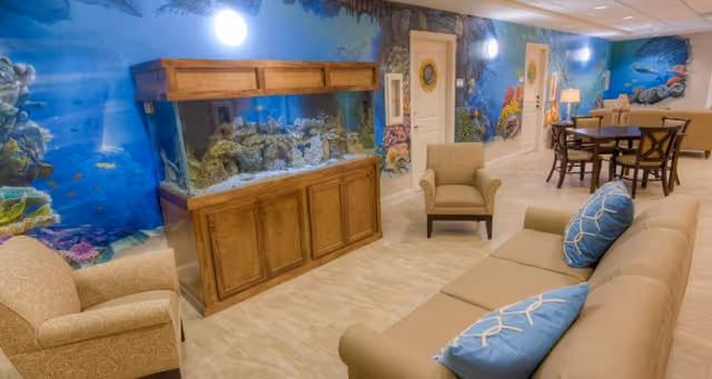 A bright communal lounge with a sofa, armchairs, an aquarium set into a wooden cabinet, an underwater mural on the wall, and a dining table in the background.