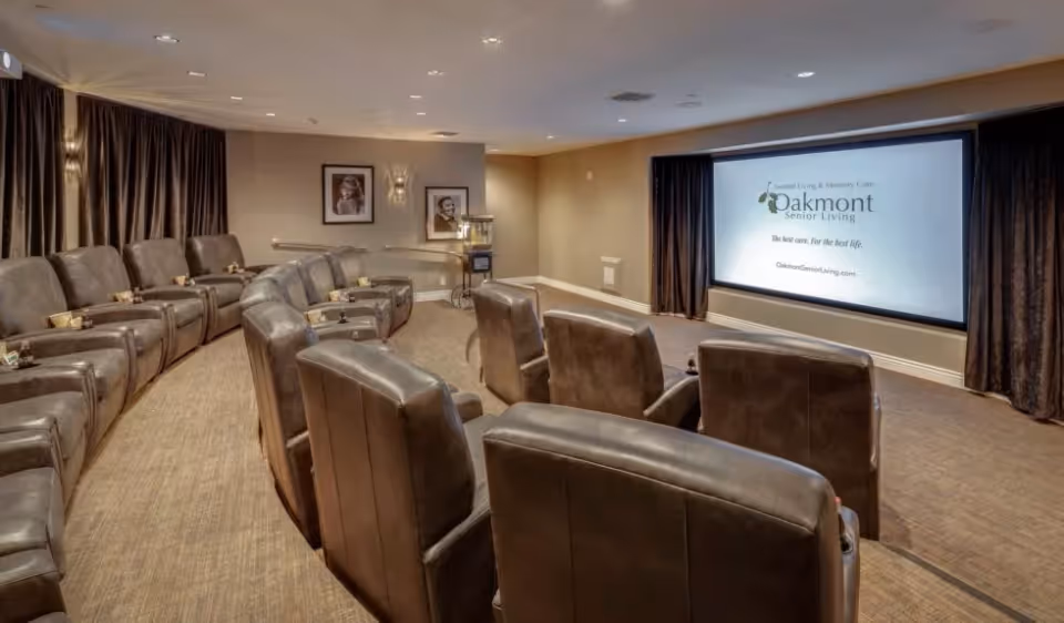 A cozy senior living facility theater room with two rows of comfortable brown leather recliners facing a large projection screen displaying the Oakmont Senior Living logo and website. The room has beige walls, carpeted floor, framed pictures on the wall, and dark curtains on the windows.