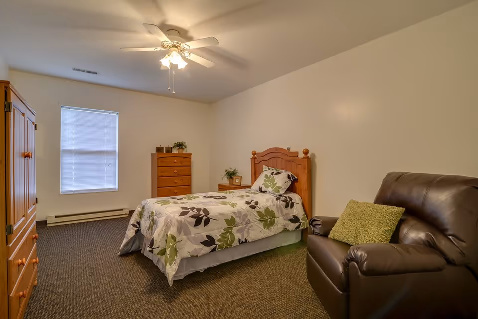 A simple bedroom with a single bed covered in a floral patterned bedspread, a wooden headboard, a wooden dresser, a wooden wardrobe, a small wooden nightstand with a plant and picture frame, a brown leather armchair with a green cushion, and a ceiling fan with lights. The room has beige walls and a window with closed blinds.