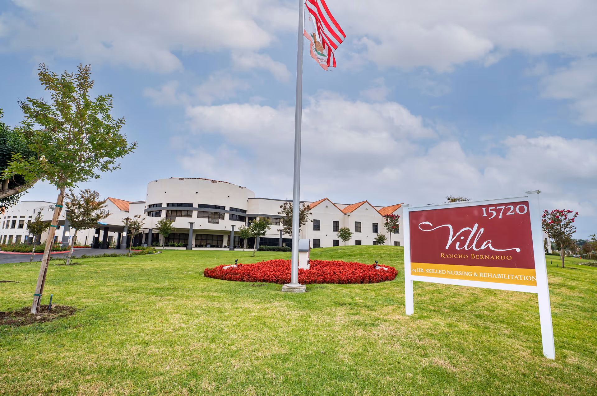 Exterior view of Villa Rancho Bernardo Care Center showing a large green lawn with a circular flower bed around a flagpole flying the American and California state flags. The building is white with a curved section and red-tiled roofs under a partly cloudy sky. A sign in the foreground reads 'Villa Rancho Bernardo 15720 24 HR. SKILLED NURSING & REHABILITATION'.