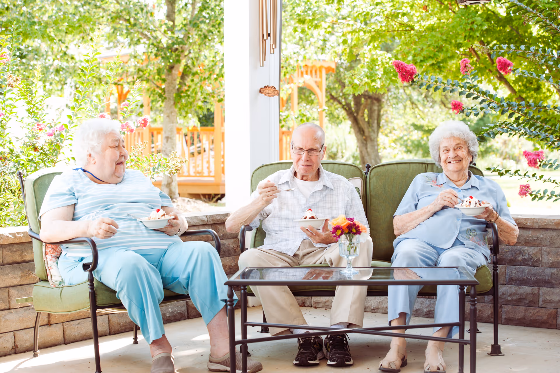 Three elderly individuals sitting on green cushioned outdoor chairs around a glass coffee table, enjoying bowls of ice cream with whipped cream and cherries. They are on a covered patio surrounded by greenery and flowering plants, with a wooden structure visible in the background.