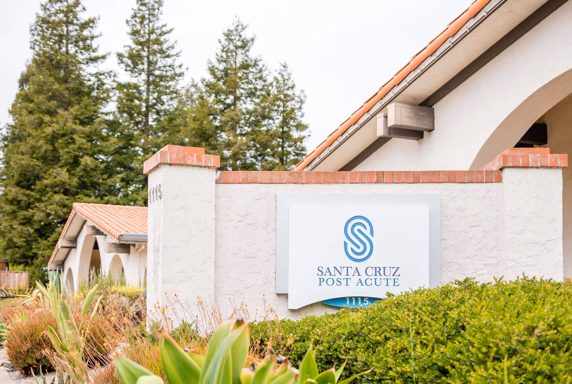 Exterior view of Santa Cruz Post Acute facility with a white stucco wall and a sign displaying the facility name and logo. The building has a tiled roof and is surrounded by green shrubs and trees.