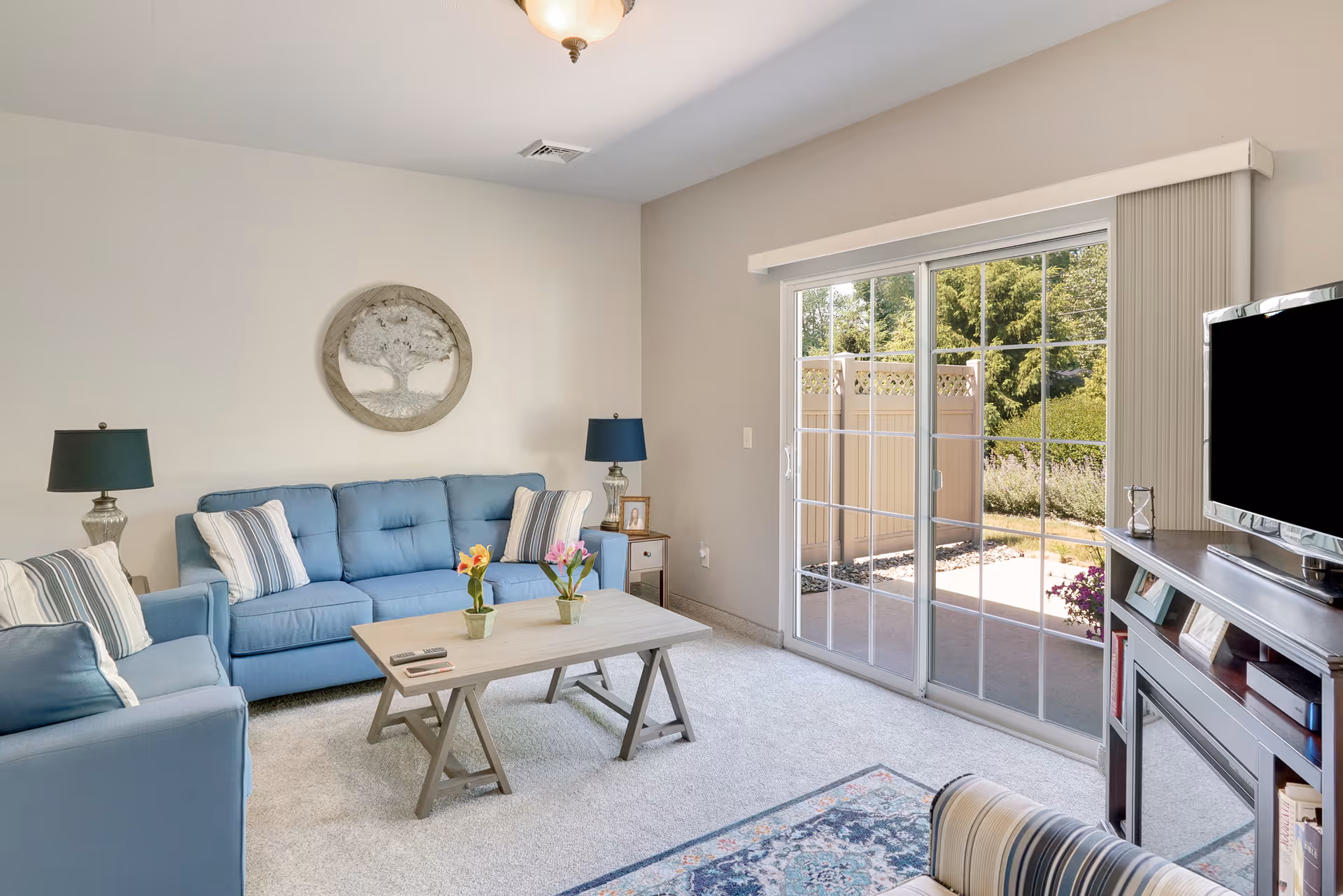 A bright living room with light blue sofas, a wooden coffee table with two small flower pots, a TV on a wooden stand with a fireplace, and a sliding glass door leading to an outdoor patio with greenery visible outside.
