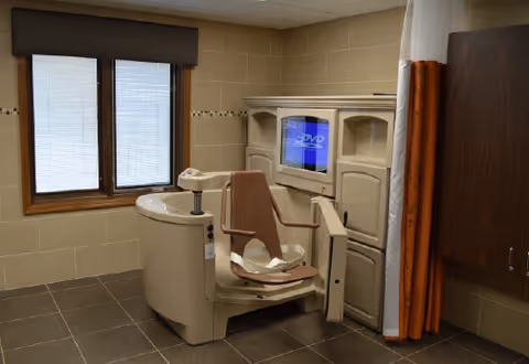 A bathroom with a specialized bathing chair and a TV mounted on a wall.