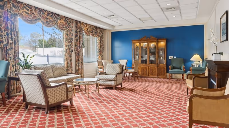 A spacious living room area with patterned red carpet, floral curtains, and large windows letting in natural light. The room features multiple upholstered chairs and sofas arranged around a glass coffee table, a wooden china cabinet against a blue accent wall, and a small table with a lamp and decorative items.