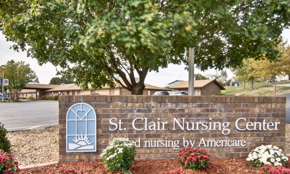 Brick sign for St. Clair Nursing Center with a logo featuring a heart and sun design, surrounded by landscaping with flowers and trees, with the nursing center building and parking lot visible in the background.