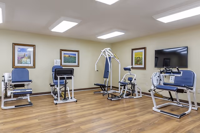 A small fitness room with four pieces of exercise equipment arranged around the room. The room has wood flooring, beige walls, three framed landscape paintings, and a flat-screen TV mounted on the wall.