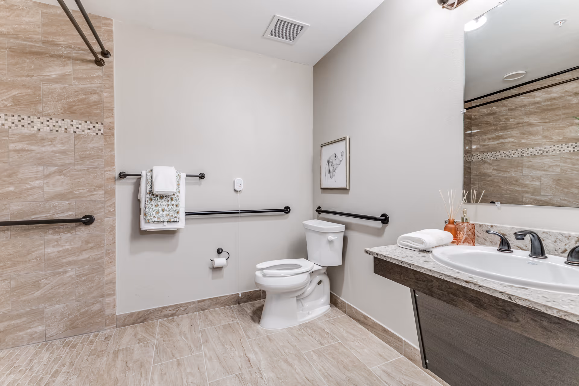 A clean and modern bathroom with beige tiled floors and walls. The bathroom features a white toilet with black safety grab bars on the walls around it. There is a towel rack with folded towels, a large mirror above a granite countertop with a white sink and black faucet fixtures. The shower area has matching beige tiles with a decorative tile border and a black grab bar.