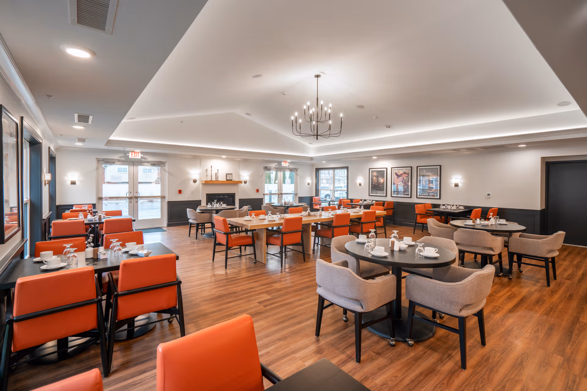 A spacious dining room with wooden floors, multiple tables set with white cups, saucers, and glasses. The room features orange and beige chairs, a modern chandelier hanging from a high ceiling, and large windows letting in natural light. There are framed pictures on the walls and a fireplace in the background.