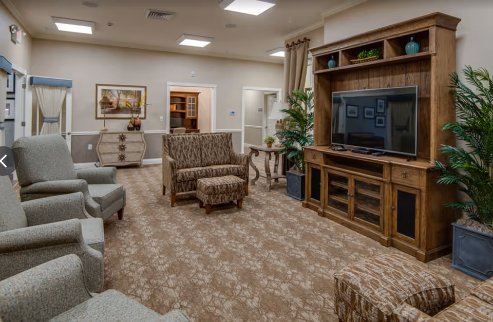 Comfortable senior living common room with upholstered chairs and sofa, a large wooden entertainment center with TV, and decorative plants.