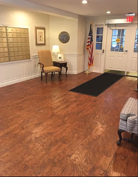 A senior living facility lobby with wood floors, mailboxes, seating, an American flag, and double entry doors.
