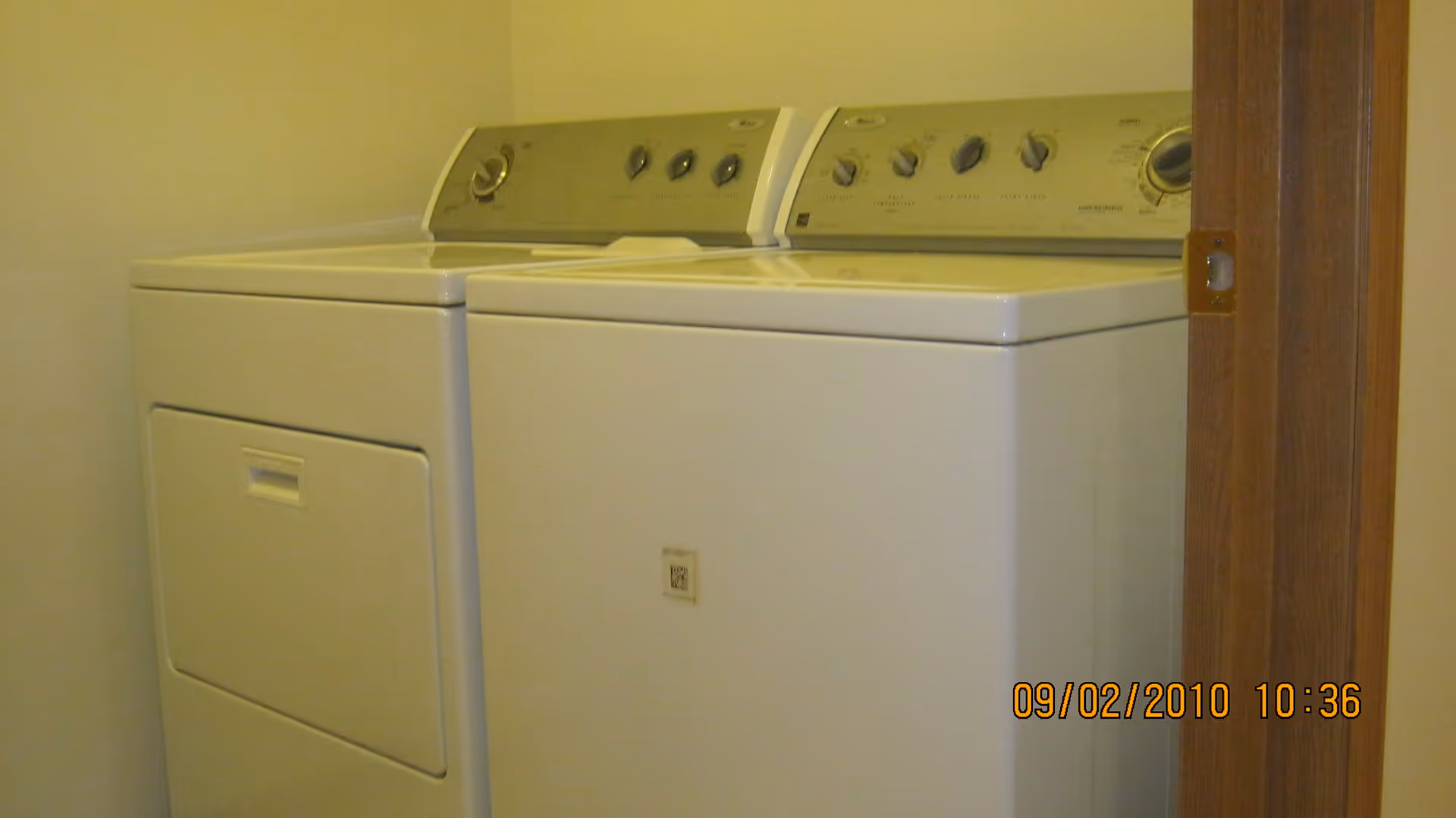 A laundry room with a white washing machine and dryer side by side against a beige wall, with a wooden door partially visible on the right.