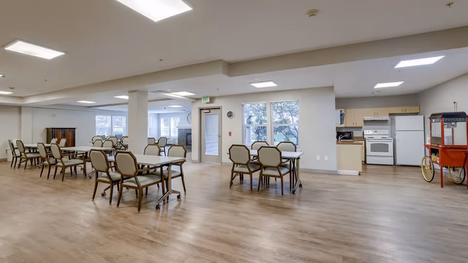 A spacious dining area with multiple tables and chairs arranged neatly on a wooden floor. The room has large windows letting in natural light, a door leading outside, and a kitchen area with a stove, refrigerator, and cabinets. A popcorn machine is visible on the right side of the room.