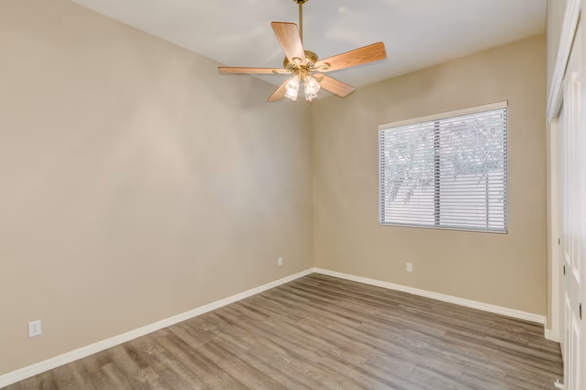 Empty bedroom with wood-look flooring, beige walls, a ceiling fan, and a window with blinds.
