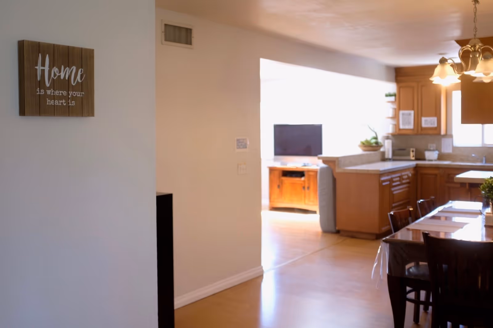 Interior view of a home showing a dining area with a wooden table and chairs, a kitchen with wooden cabinets and countertops, and a living room area with a TV on a wooden stand. A decorative wooden sign on the wall reads 'Home is where your heart is.'
