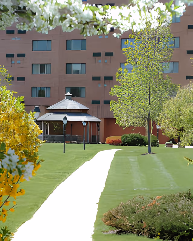 A paved white walkway leads across a green lawn to a gazebo with a multi-story brick building and windows in the background, framed by trees and shrubs.